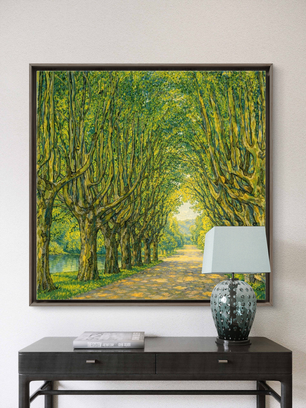A framed landscape painting of a sunlit tree-lined path with green foliage, displayed on a light wall above a dark console table with a lamp and book.
