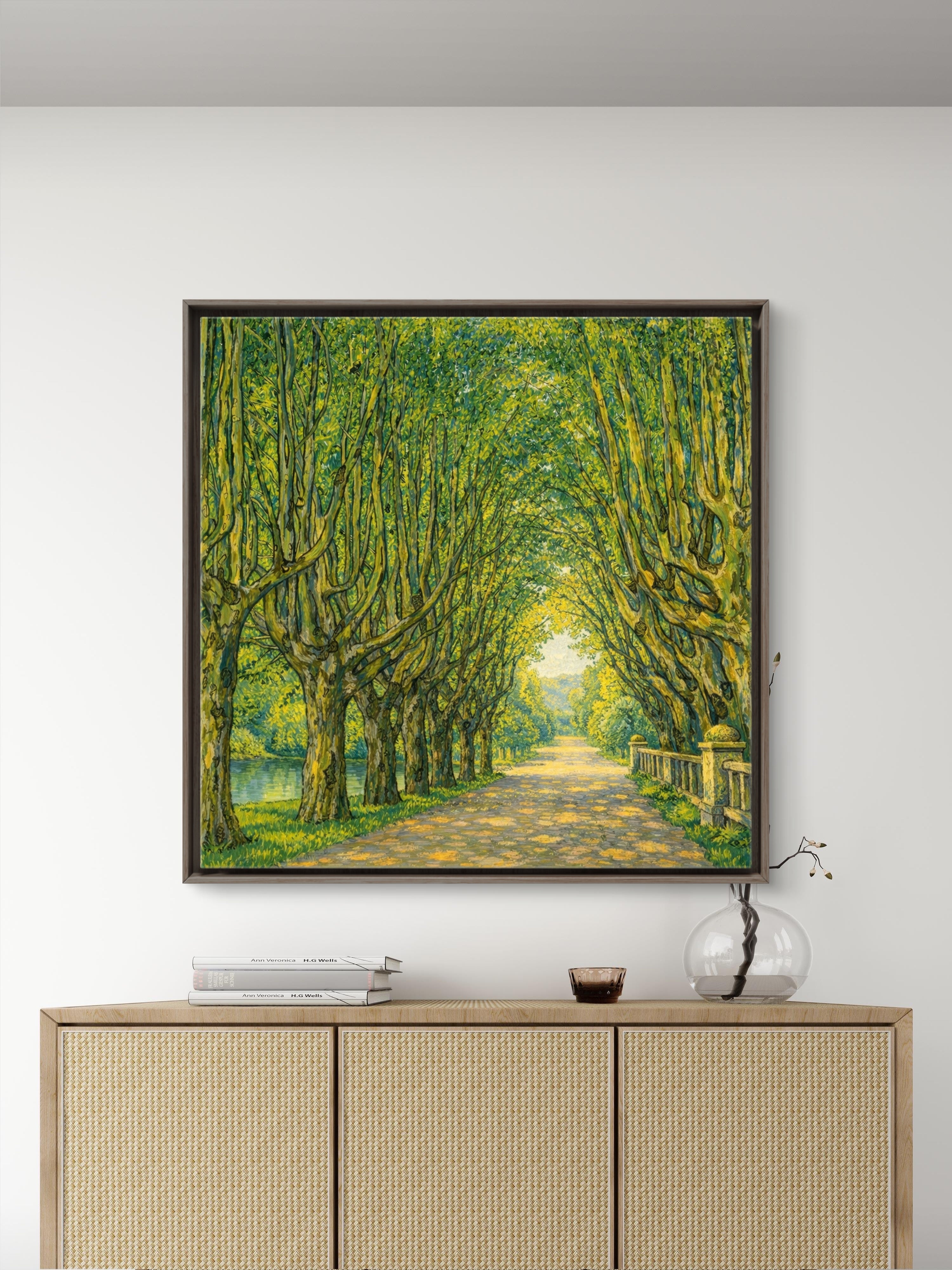 A framed impressionist-style painting of a sunlit tree-lined path hangs on a white wall above a light wood cabinet with woven panels and small decor objects on top.