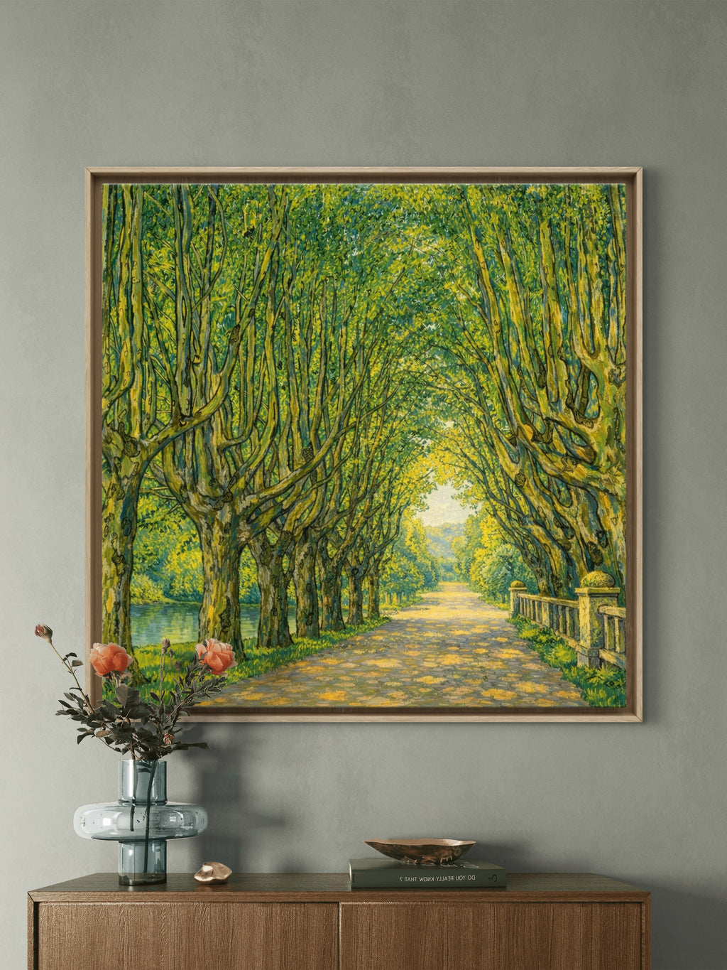 A framed landscape painting of a sunlit tree-lined path hangs on a gray wall above a wooden console table with a glass vase of flowers and decorative objects.
